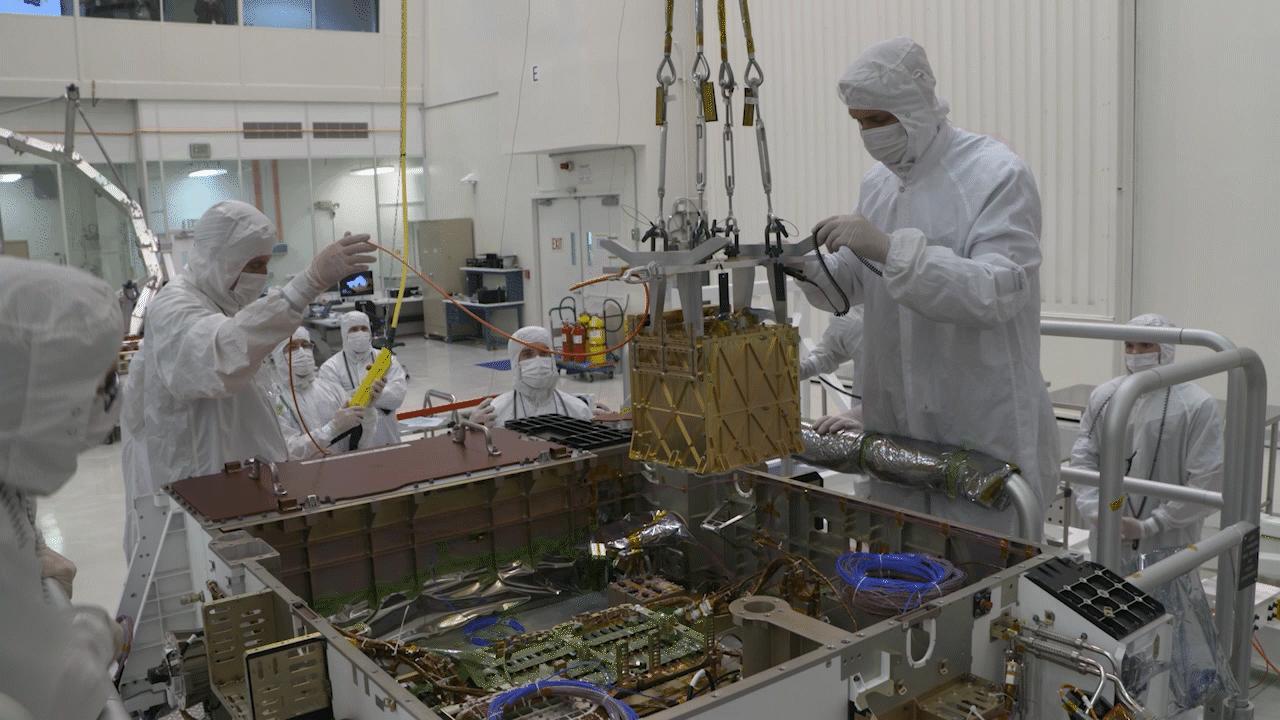 Engineers in clean-room suits lower the gold-coloured MOXIE instrument into NASA’s Perseverance rover